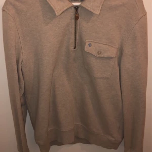 Beige tröja med dragkedja från Morris - Säljer en stilren beige tröja från Morris med dragkedja och krage. Tröjan har en bröstficka med knapp och en liten blå broderad detalj. Perfekt för en avslappnad men snygg look.