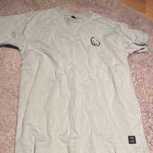 Ljusgrön t-shirt med smiley från Lager 157 - Säljer en ljusgrön t-shirt från Lager 157 med en svart smiley på bröstet. T-shirten är kortärmad och gjord i mjukt bomullsmaterial. Perfekt för en avslappnad stil.