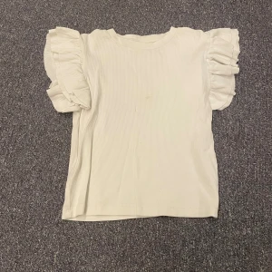 Vit ribbad topp från Gina Tricot - Säljer en söt vit ribbad topp från Gina Tricot med volangdetaljer på ärmarna. Perfekt för en stilren och trendig look. Toppen har en normal passform och är tillverkad i ett mjukt material. Har en liten fläck runt magen.