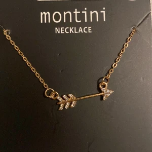 Guldhalsband med pil från Montini - Elegant guldhalsband från Montini med en pilformad berlock dekorerad med glittriga stenar. Perfekt för att ge en stilren touch till din outfit. Kedjan är tunn och smidig, vilket gör den lätt att bära.