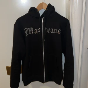 Svart hoodie från Masteme - Säljer en svart hoodie från Masteme med dragkedja och ett coolt tryck på baksidan. Framsidan har text i glittrig stil. Perfekt för dig som vill ha en unik stil.