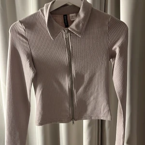 Beige tröja - Säljer en beige ribbad jacka med dragkedja framtill. Jackan har en klassisk krage och långa ärmar. Perfekt för en stilren look.