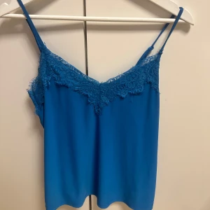 Blå linne med spetsdetaljer - Snygg blå linne med smala axelband och vacker spetsdetalj längs halsringningen. Perfekt för varma dagar eller som ett lager under en kavaj. Enkel och stilren design som passar till många olika outfits.