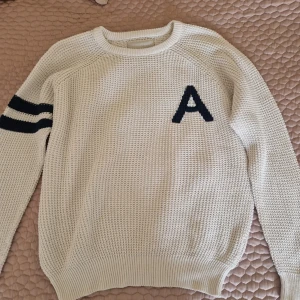 Vit stickad tröja från Iconic Sport - Säljer en vit stickad tröja från Iconic Sport med ett stort svart 'A' på bröstet och två svarta ränder på ena ärmen. Tröjan har en rund halsringning och ribbstickade muddar vid ärmslut och nederkant. Perfekt för en sportig look!