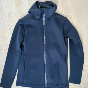 Mörkblå huvtröja från J.Lindeberg - Snygg mörkblå huvtröja från J.Lindeberg med dragkedja framtill. Tröjan har en stilren design med hög krage och långa ärmar. Perfekt för en modern och sportig look. Storleken är large men är mer som storlek medium 