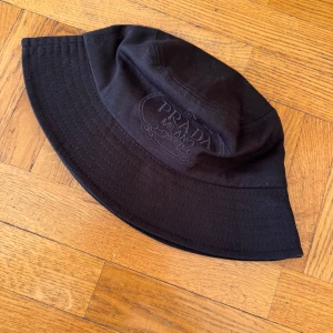 Svart bucket hat från Prada - Snygg svart hatt från Prada med broderad logotyp. Perfekt för en stilren look. Tillverkad i ett slitstarkt material.