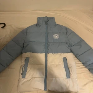 Blå och vit pufferjacka från Supply & Demand - Säljer en snygg blå och vit pufferjacka från Supply & Demand. Jackan har en dragkedja framtill och ett coolt märke på bröstet. Perfekt för kyliga dagar 