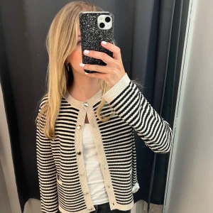 Randig kofta i svart och beige - Säljer en snygg randig kofta i svart och beige med knappar framtill. Koftan har långa ärmar och en klassisk rund halsringning. Perfekt för en stilren look. Nypris 600kr, prislapp kvar