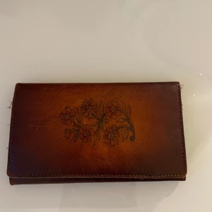 Brun skinnplånbok med blommönster - Säljer en stilren brun skinnplånbok med ett vackert blommönster på framsidan. Plånboken har flera fack för kort och en transparent ficka för ID. Perfekt för att hålla ordning på dina tillhörigheter.Brunt persisk handgjord plånbok i äkta läder . Med fem fickor . Bred :16,05cm  Längd :9,08cm ( som är stängd) Längd :ca22cm ( öppet dom på bild tre)  Har köpt år 2000