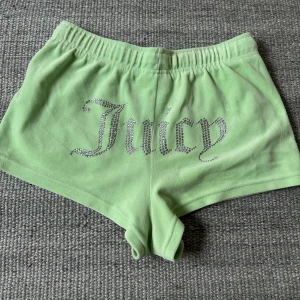 Gröna shorts från Juicy Couture storlek S - Snygga ljusgröna shorts från Juicy Couture med glittrande text på baksidan. De har en bekväm resårmidja och snörning framtill samt praktiska dragkedjefickor. Perfekta för en avslappnad stil.