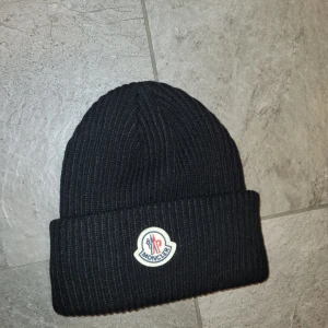 Svart moncler mössa - Snygg svart ribbad mössa från Moncler med broderad logotyp framtill. Perfekt för att hålla värmen under kyliga dagar. Stilren design som passar till det mesta. Inga byten.  Priset kan diskuteras. 