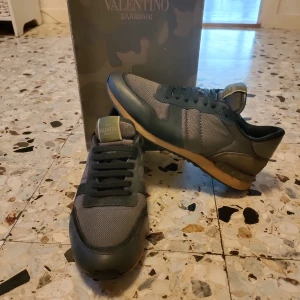 Valentino rockrunners - Snygga Valentino Garavani rockrunners sneakers i grått med svarta detaljer och en beige sula. Skorna har en stilren design med snörning och är tillverkade i en kombination av material för en modern look. Skick 8/10 på övre delen av skon är de 9.5/10 skick ser nästan helt oanvända ut det är bara sulan som har slipats ner lite men man märker det inte mycket 