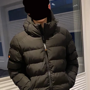 Svart pufferjacka från Superdry - Säljer en svart pufferjacka från Superdry med dragkedja och hög krage. Jackan har en vadderad design och långa ärmar, perfekt för kyliga dagar. Den har en logotyp på ärmen och en justerbar huva för extra skydd mot väder och vind. Köpt från Stadium och använts endast ett fåtal gånger. Köptes i December och är som ny! 490kr för denna men 790kr vid köp av båda jackorna i min annons.