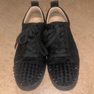 Svarta Christian Louboutin skor - Snygga svarta sneakers i mocka från Christian Louboutin med ikoniska röda sulor. Skorna har svarta nitar på tån och snörning framtill. Perfekta för att ge en edgy touch till din stil.