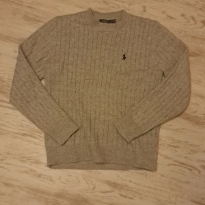 Beige stickad tröja från Polo Ralph Lauren - Säljer en stilren beige stickad tröja från Polo Ralph Lauren med kabelstickat mönster. Tröjan har långa ärmar och en liten broderad logotyp på bröstet. Perfekt för kyliga dagar.