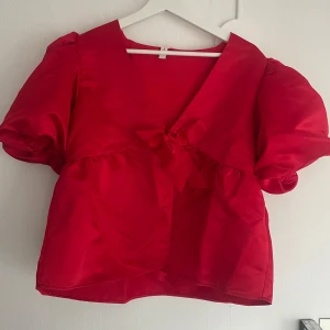Röd blus med puffärmar - Snygg röd blus med puffiga kortärmar och en söt rosett framtill. Blusen har en v-ringad design och är tillverkad i ett glansigt material som ger en lyxig känsla. Använd 1 gång!! 💞💞