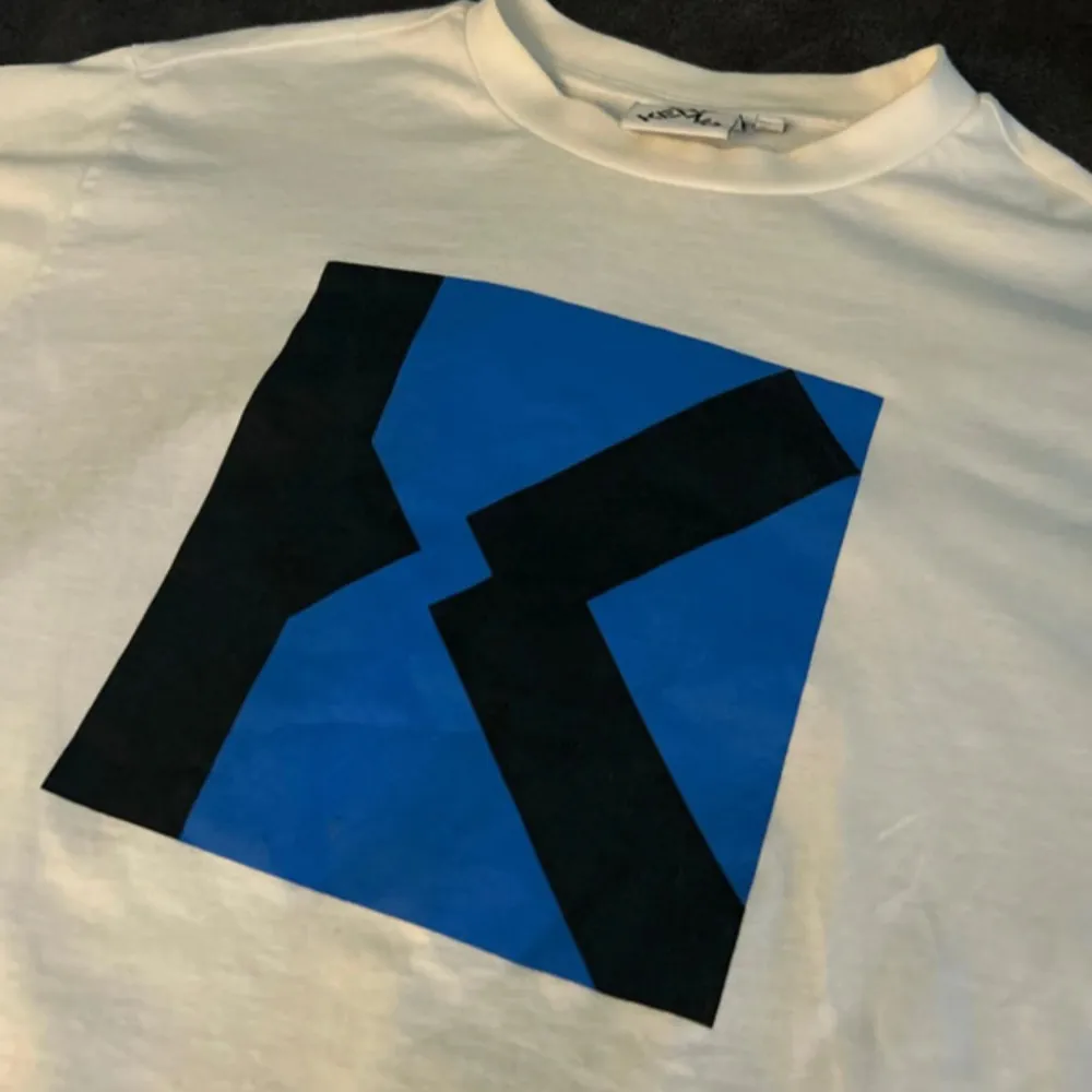 Säljer en stilren vit t-shirt från Kenzo med ett iögonfallande blått och svart grafiskt tryck på framsidan. Perfekt för dig som vill ha en unik och trendig look. T-shirten är kortärmad och tillverkad i mjukt material för extra komfort.. T-paidat.
