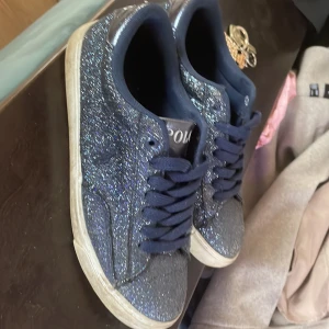 Glittriga sneakers från ralp loren - Snygga glittriga sneakers från ralp loren i en mörkblå nyans. Skorna har snörning och en vit sula som ger en cool kontrast. Perfekta för att lägga till lite glitter i vardagen. Ska tvätta dom innan 🥰