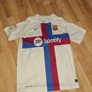 Vit FC Barcelona fotbollströja - Säljer en vit FC Barcelona fotbollströja från Nike med ett stort kors i blått och rött på framsidan. Tröjan har korta ärmar och La Liga-logga på ärmen. Perfekt för fans av laget!