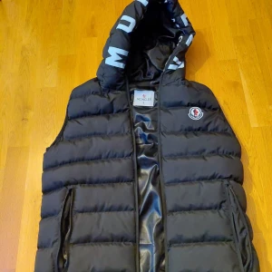 Svart dunväst från Moncler - Snygg svart dunväst från Moncler med huva och dragkedja. Västen har Moncler-logga på bröstet och tryck på insidan av huvan. Perfekt för kyligare dagar, är äkta också.