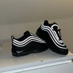 Svarta Nike Air Max sneakers - Snygga svarta Nike Air Max sneakers med reflekterande detaljer. Skorna har en ikonisk design med synlig luftdämpning i sulan och snörning framtill. Perfekta för en stilren look.HELT NYA 