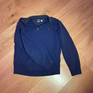 Mörkblå tröja från Polo Ralph Lauren - Säljer en stilren mörkblå tröja från Polo Ralph Lauren med dragkedja vid halsen. Tröjan har långa ärmar och en liten röd logotyp på bröstet. Perfekt för en avslappnad och snygg look.