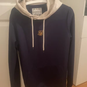 Mörkblå hoodie från SikSilk - Snygg mörkblå hoodie från SikSilk med beige huva och dragsko. Tröjan har en diskret logga på bröstet och dragkedja på sidorna. Fint skick! 