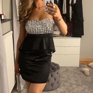 Glittrig axelbandslös klänning - Snygg axelbandslös klänning med en glittrig silveröverdel och svart kjol. Klänningen har en volangdetalj i midjan som ger en elegant touch. Perfekt för festliga tillfällen💖