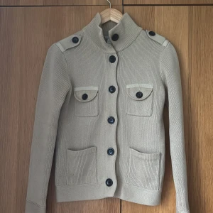 Stickad Beige kofta från Filippa K - Säljer en stilren beige kofta från Filippa K i 100% bomull. Koftan har långa ärmar och knappar framtill samt fyra fickor med knappdetaljer. Perfekt för en elegant och avslappnad look. (Nyskick)