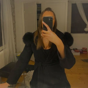 Svart kappa med pälskrage - Elegant svart kappa med stor pälskrage och långa ärmar. Perfekt för kyliga dagar med sin stilrena design och bekväma passform. Ordinarie pris 5000kr.