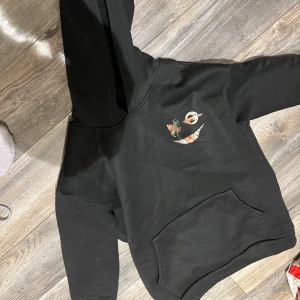 Svart hoodie med tryck - Cool svart hoodie med unikt tryck i orange och vitt på både fram- och baksidan. Perfekt för en avslappnad stil med en touch av edge. Hoodien har en klassisk känguruficka och en bekväm huva. En cool hoodie som värmer dig i vinter! Snörena på luvan är av. 