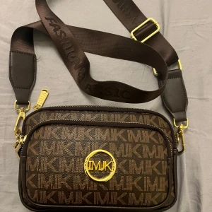 Brun axelväska  - Snygg brun axelväska från  med guldiga detaljer och logotyp. Väskan har ett justerbart axelband och flera dragkedjefack för praktisk förvaring. Perfekt för att bära med sig det viktigaste med stil.