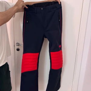 Mörkblåa och röda skidbyxor med dragkedjor - Snygga svarta byxor med röda detaljer och dragkedjor på sidorna. Perfekta för en sportig look. Byxorna har en modern design med quiltade knäpartier och en bekväm passform. Oanvänd med prislapp. Säljer matchande skidjacka