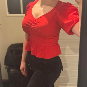 Röd topp med volangdetalj - Snygg röd topp med volangdetalj och puffiga kortärmar. Toppen har en v-ringad design och rynkade detaljer framtill som ger en framhävande effekt. Perfekt för tex julfirande. Säljer pga att den är förliten för mig! Toppen är i S men upplever den stor i storleken så tror den passar M också. Ny med lapp kvar.