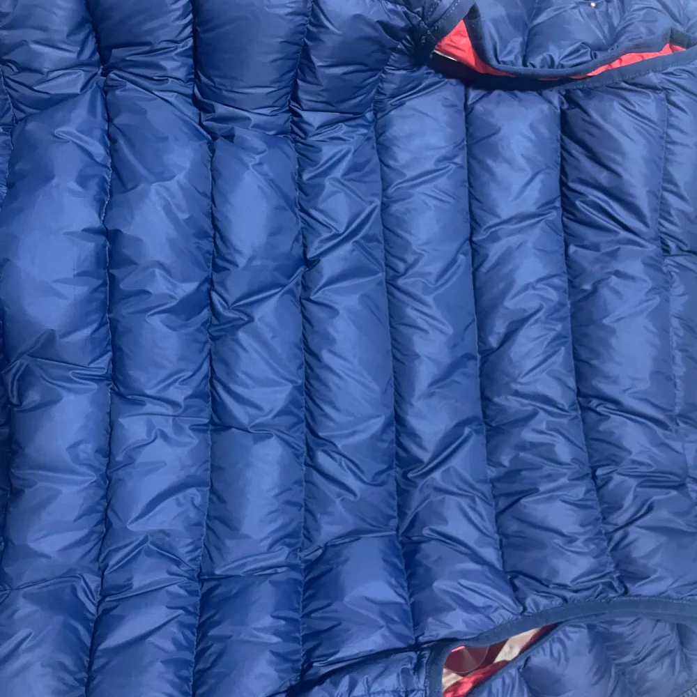 Snygg och varm dunväst från Tommy Hilfiger i blått med röd insida. Västen har en dragkedja framtill och är perfekt för kyligare dagar. Den quiltade designen ger en stilren look.. Takit.