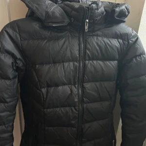 Svart dunjacka från Rockandblue - Snygg svart dunjacka från Rockandblue med dragkedja och huva. Den har en figurnära passform och sitter som en smäck! 