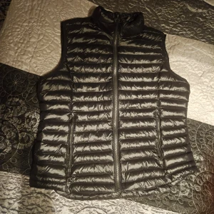 Svart dunväst från Everest - Säljer en stilren svart dunväst från Everest. Västen har en dragkedja framtill och är perfekt för kyligare dagar. Den är lätt och smidig att bära, vilket gör den till ett mångsidigt plagg i garderoben.