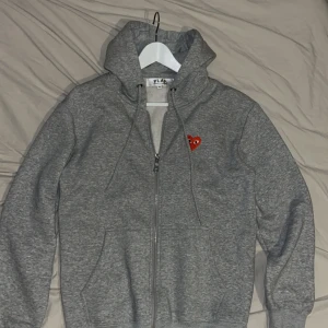 Grå zip hoodie från Comme des Garcons Play - Säljer en stilren grå hoodie från Comme des Garçons Play med dragkedja och det ikoniska röda hjärta-emblemet på bröstet. Perfekt för en casual look med en touch av design. Tröjan är också otroligt skön. 