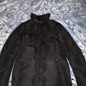 Svart pufferjacka från Hollies - Snygg svart pufferjacka från Hollies med camouflagemönstrat foder och orange detaljer. Jackan har flera praktiska fickor och en hög krage för extra värme. Perfekt för kyliga dagar!