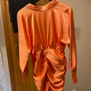 Orange sidenklänning från C/Meo - Snygg orange klänning i siden från C/Meo med långa ärmar och en elegant rynkad detalj på baksidan. Klänningen har en hög krage och en öppen rygg som ger en stilren look. Perfekt för att sticka ut med sin glansiga finish.