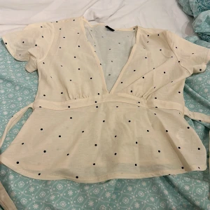 Kräm Vit blus med prickar - Gina Tricot Tie Top  - Kräm vit blus med små svarta prickar och v-ringning. Blusen har korta ärmar och knytband i midjan för en fin passform. Sparsamt använd. Inga defekter. 