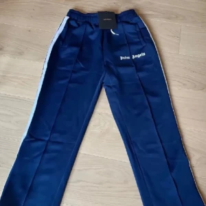 Blå träningsbyxor från Palm Angels - Snygga blå träningsbyxor från Palm Angels med vita sidoremsor och logotyp på benet. Byxorna har en normal passform och är perfekta för en sportig look.