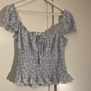 Blommig topp från Gina Tricot - Söt blommig topp från Gina Tricot i ljusblått och vitt. Toppen har korta puffärmar och en smockad midja med volangdetaljer. 