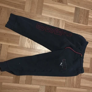 Svarta mjukisbyxor från Puma  - Säljer ett par svarta mjukisbyxor från  Puma med röd text och logga på sidan. Byxorna har en bekväm passform med resår i midjan och vid bensluten. Perfekta för en avslappnad stil.