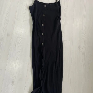 Svart långklänning med knappar - Elegant svart långklänning med knappar framtill. Klänningen har smala axelband och en stilren design som passar perfekt för olika tillfällen.