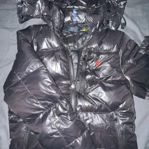Svart pufferjacka från Polo Ralph Lauren - Snygg svart pufferjacka från Polo Ralph Lauren med glansig finish. Jackan har en praktisk dragkedja och en huva för extra skydd mot väder. Perfekt för kyliga dagar.