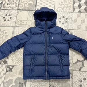 Blå dunjacka från Polo Ralph Lauren - Säljer en blå dunjacka från Polo Ralph Lauren med huva och dragkedja. Jackan har långa ärmar och två fickor med dragkedja framtill. Perfekt för kyliga dagar.