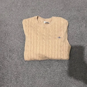 Beige stickad tröja från GANT - Säljer en stilren beige stickad tröja från GANT med klassiskt kabelstickat mönster. Perfekt för kyligare dagar och ger en tidlös look. Tröjan har rund hals och ribbade kanter.
