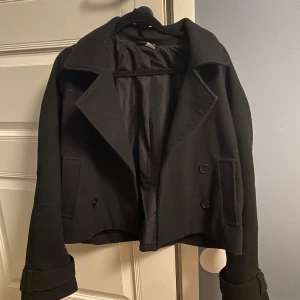 Svart kappa med knappar - Snygg svart kappa med dubbelknäppning och bred krage. Jackan har långa ärmar och stilrena detaljer som knappar på axlarna. Perfekt för en elegant look.