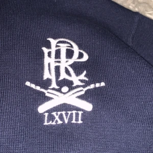 Mörkblå tröja från Polo Ralph Lauren - Säljer en mörkblå tröja från Polo Ralph Lauren med dragkedja och broderad logga på bröstet. Tröjan har långa ärmar med vita ränder på ena ärmen och en ribbad krage. Perfekt för en stilren och sportig look.